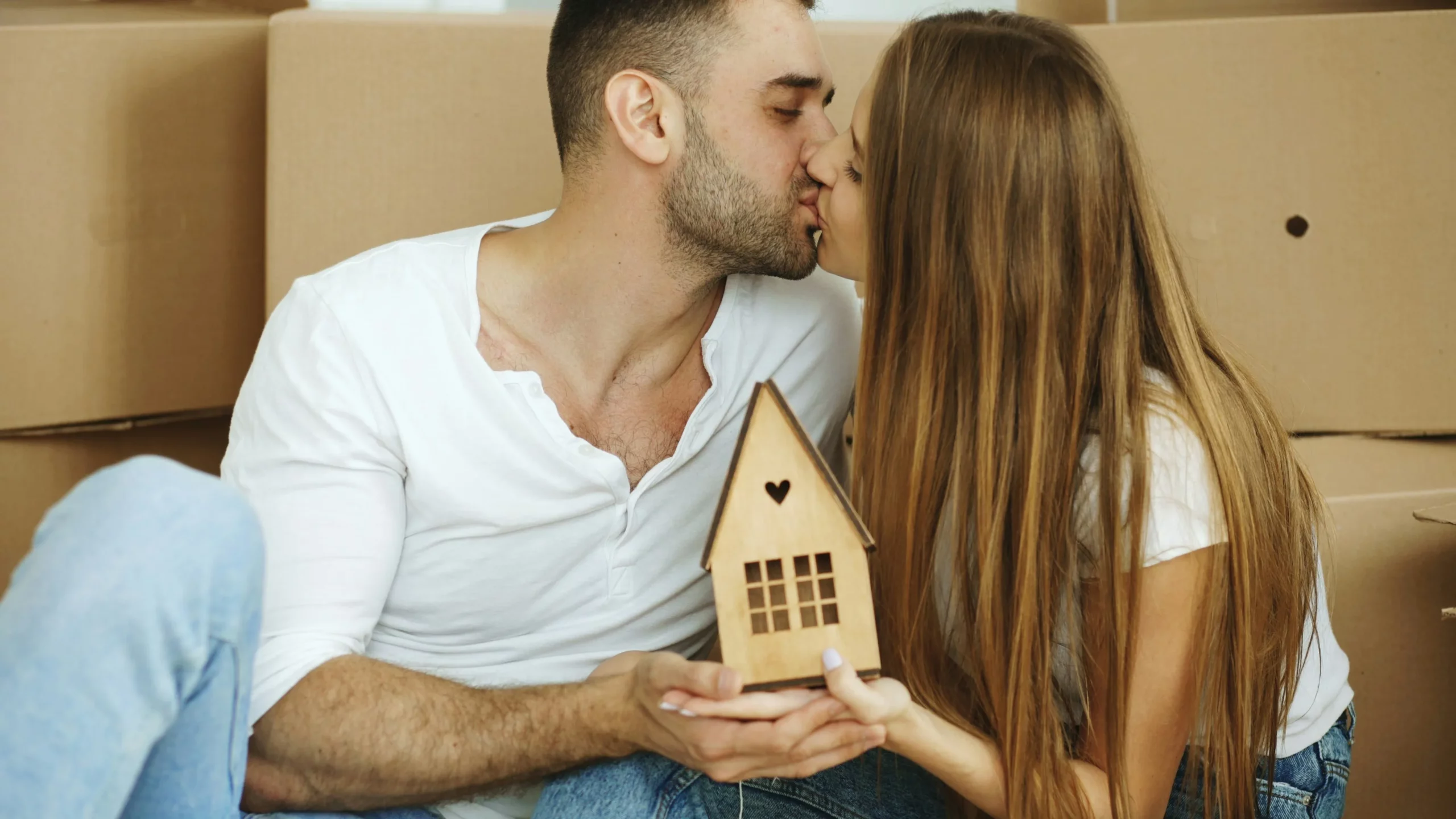 Couple s'embrassant en tenant une maison miniature en bois, cartons de déménagement